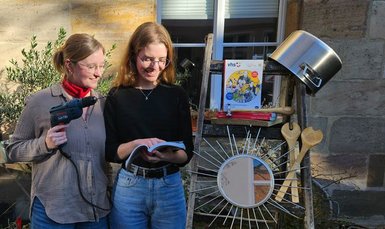 Zei Personen stehen mit dem neuen Programmheft vor einer Leiter, weiterhin auf dem Bild ein Topf, eine Bohrmaschine, Werkzeug für Schmuckbearbeitung