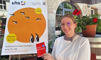 Mitarbeitende steht vor dem Plakat mit dem Semestermotiv und hält eine Karte hoch mit dem Text jetzt anmelden. Das Semestermotiv besteht aus einem orangen Kreis mit fliegenden Pinguinen denen zwei stehende Pinguine nachschauen