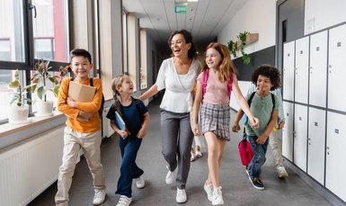 Eine Lehrkraft ist mit 4 Kindern auf einem Schulflur unterwegs, alle sind fröhlich